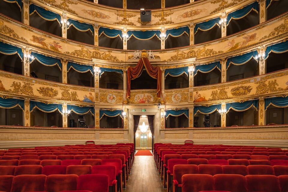 A grand theater with ornate gold and blue decorations, red velvet seats, and a central stage.