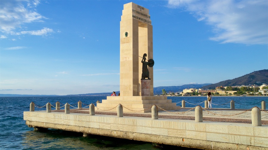 Een monument met een standbeeld op een pier aan de zee, met uitzicht op een kustplaats.
