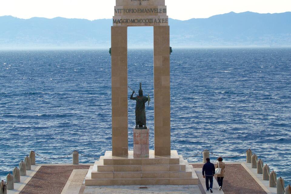 A statue of a warrior on a pedestal with inscriptions, overlooking a body of water with mountains in the background.