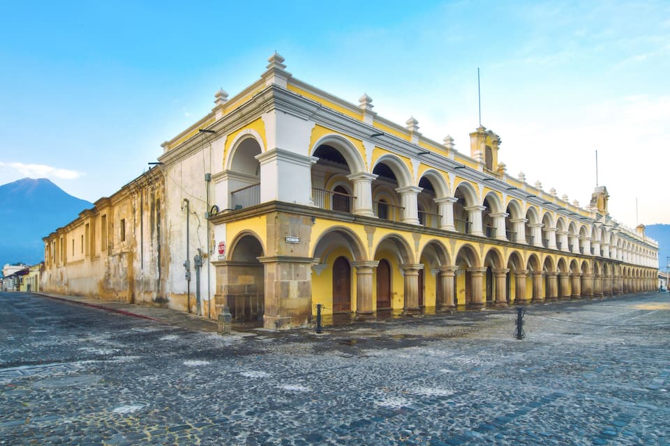 A historic building with yellow and white architecture, featuring multiple arches and columns, situated on a cobblestone street.