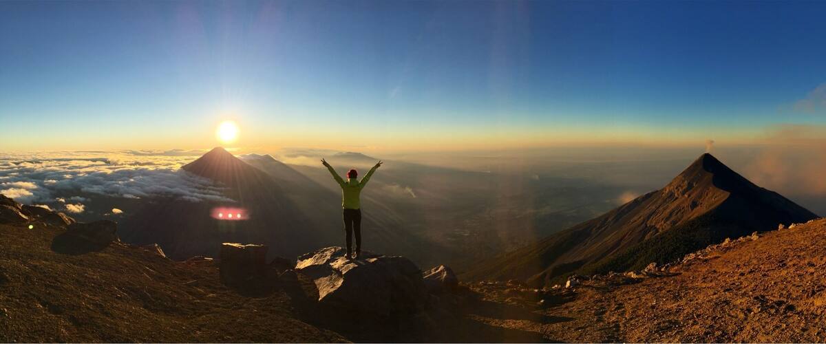 A person standing on a mountain peak with arms raised, overlooking a valley with a mountain in the distance and the sun shining through the clouds.