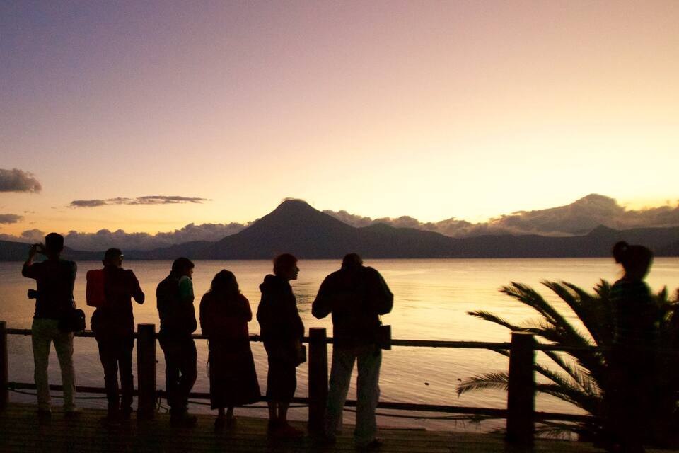 A group of people silhouetted against a sunset by a body of water with mountains in the background.