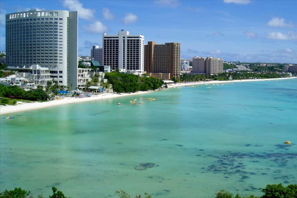 A coastal cityscape with a beach, clear turquoise waters, and high-rise buildings.