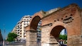 Antiguos arcos de piedra con intrincados grabados, frente a edificios modernos y un cielo azul despejado.
