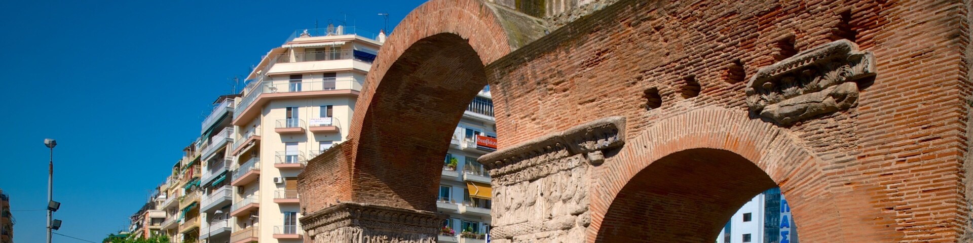 Ancient stone arches with intricate carvings, set against a backdrop of modern buildings and a clear blue sky.