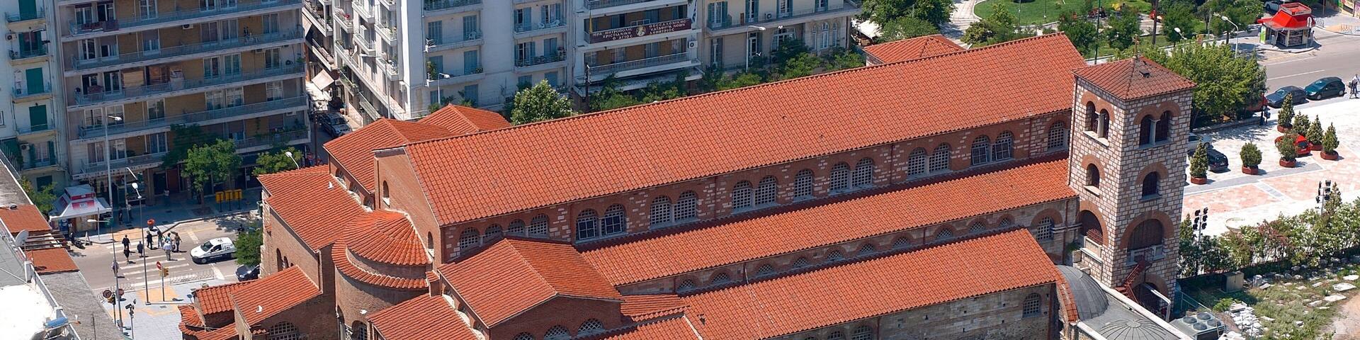 A historic building with a red-tiled roof and a dome, surrounded by modern buildings and greenery.
