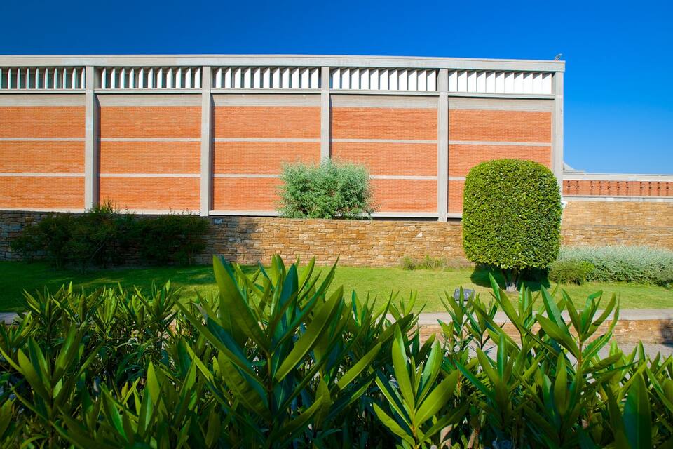 Edifici modern amb façana de maó i mur de pedra. Hi ha un jardí ben cuidat amb plantes variades i un arbust ben podat.