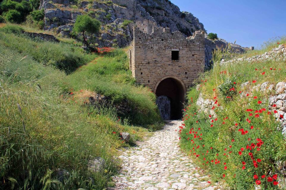 A stone pathway leading to a small stone building with an arched entrance, surrounded by greenery and red flowers.