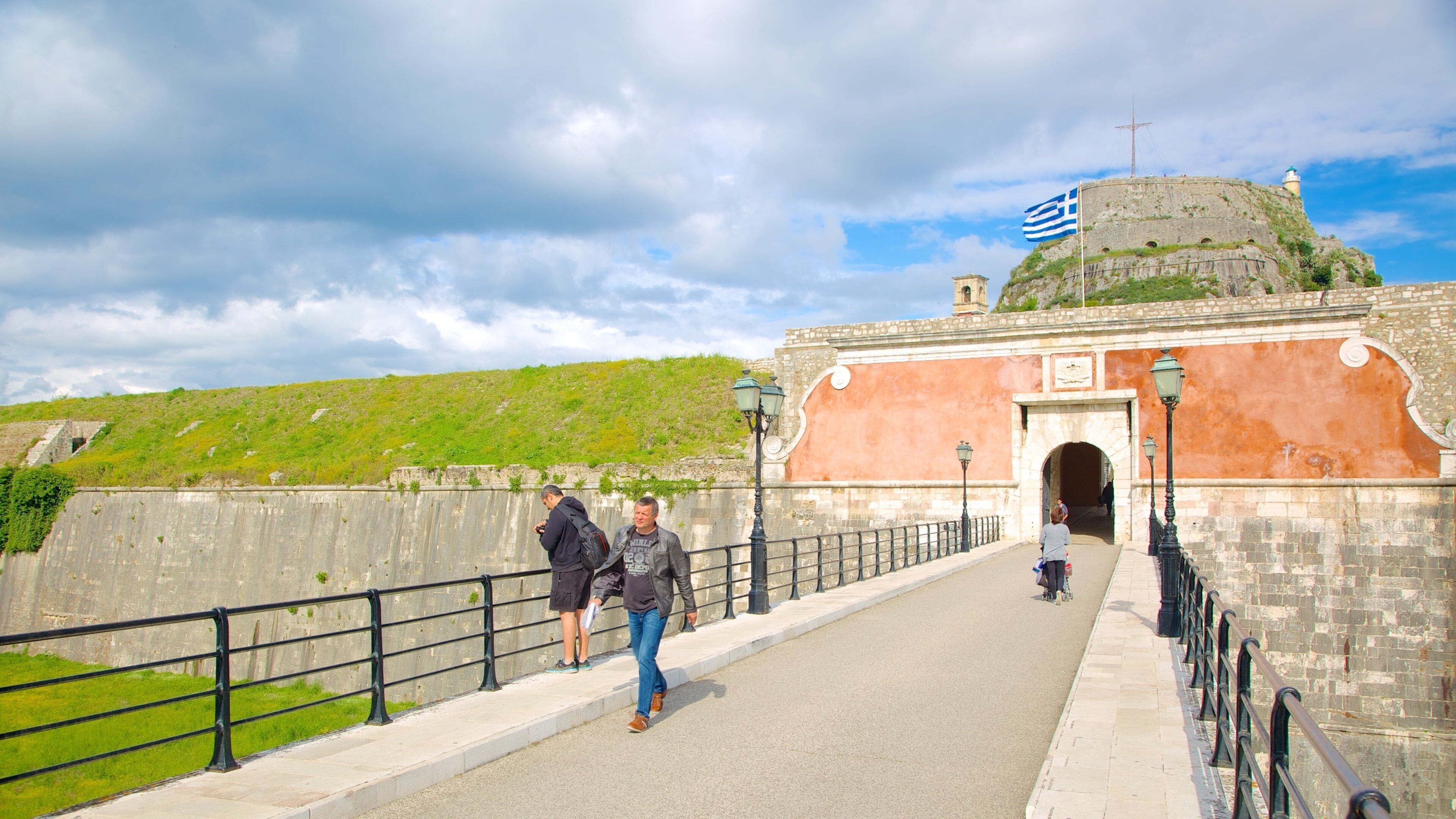 A historical fort with a flag, a paved walkway, and a green hill in the background.