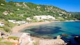 A coastal town with a sandy beach, clear blue waters, and lush green hills in the background.