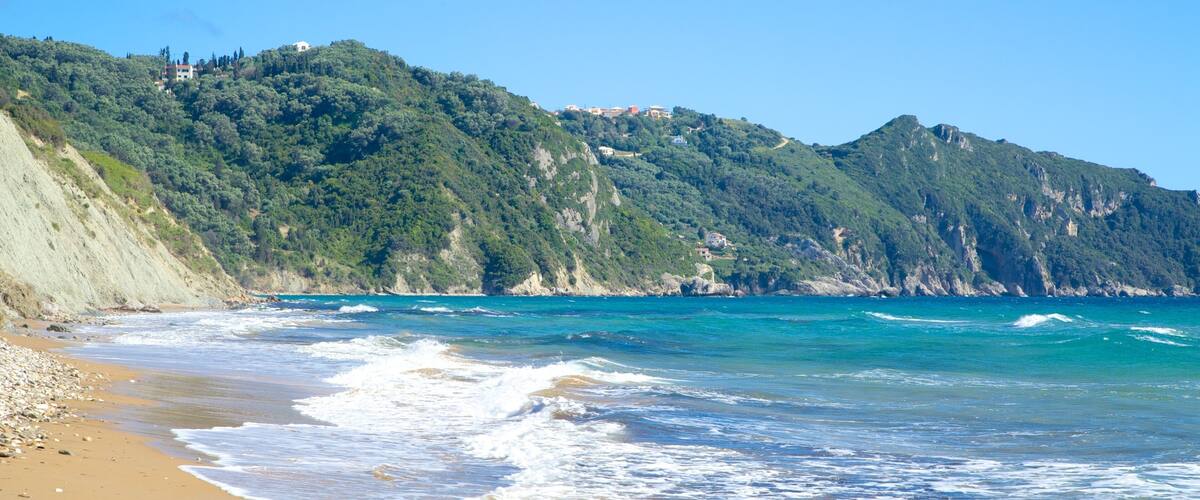 A coastal landscape with a sandy beach, turquoise sea, and green hills with buildings.