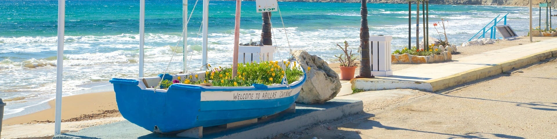 A coastal scene with a blue boat labeled "Welcome to Arillas" on the beach, a sign for the "Green Bus Time Table," and a rocky cliff in the background.