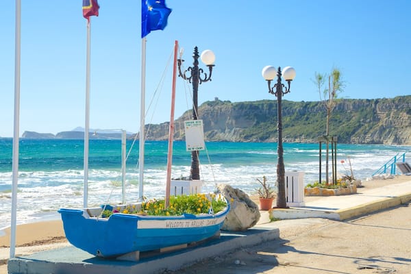 Une scène côtière avec un bateau bleu sur lequel on peut lire « Bienvenue à Arillas », une pancarte indiquant les horaires du « Green Bus » et une falaise rocheuse en arrière-plan.