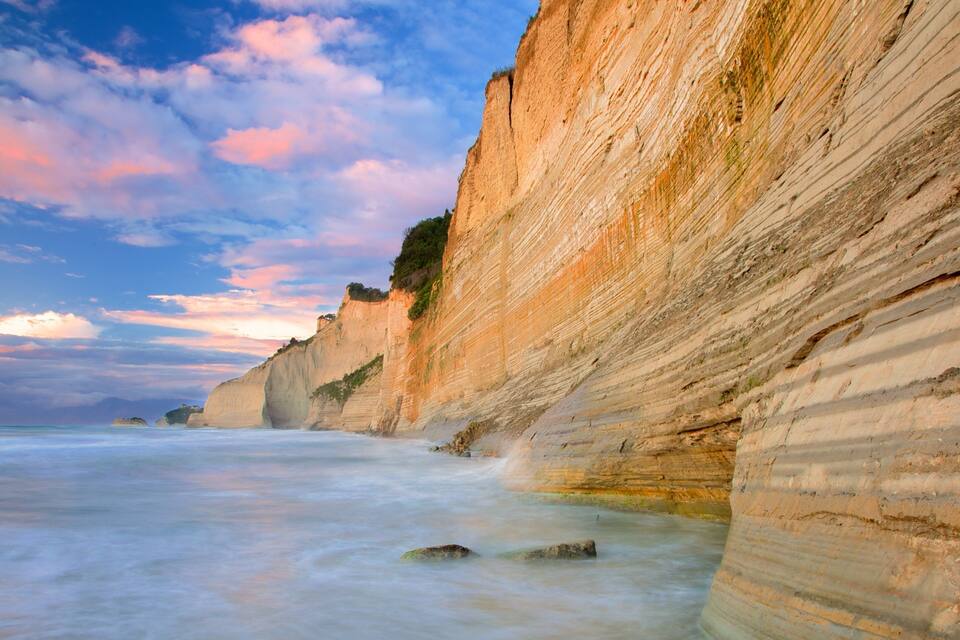 Acantilado costero con formaciones rocosas sedimentarias estratificadas y un mar tranquilo.