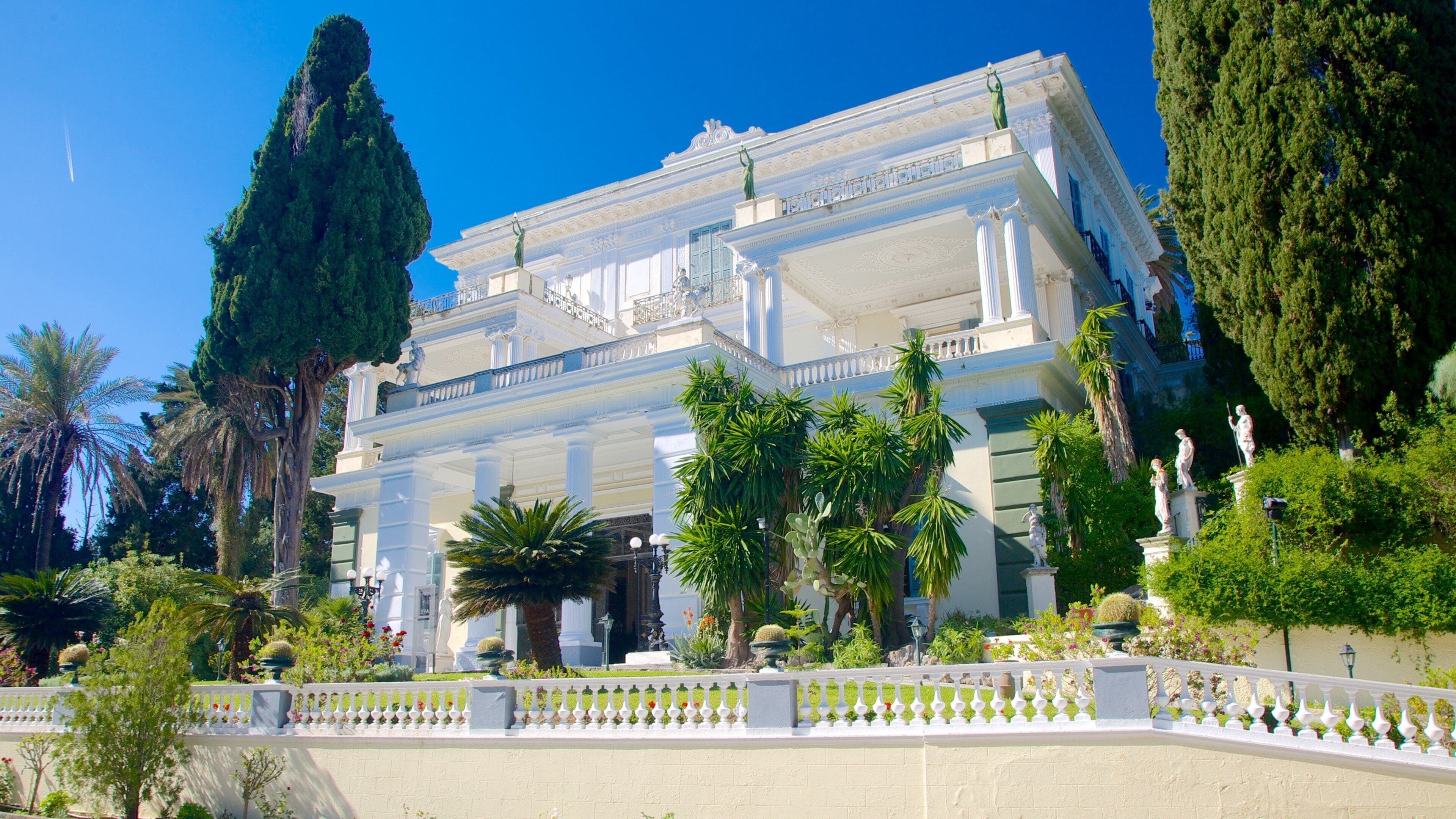 A classical building with columns, statues, and a white fence.