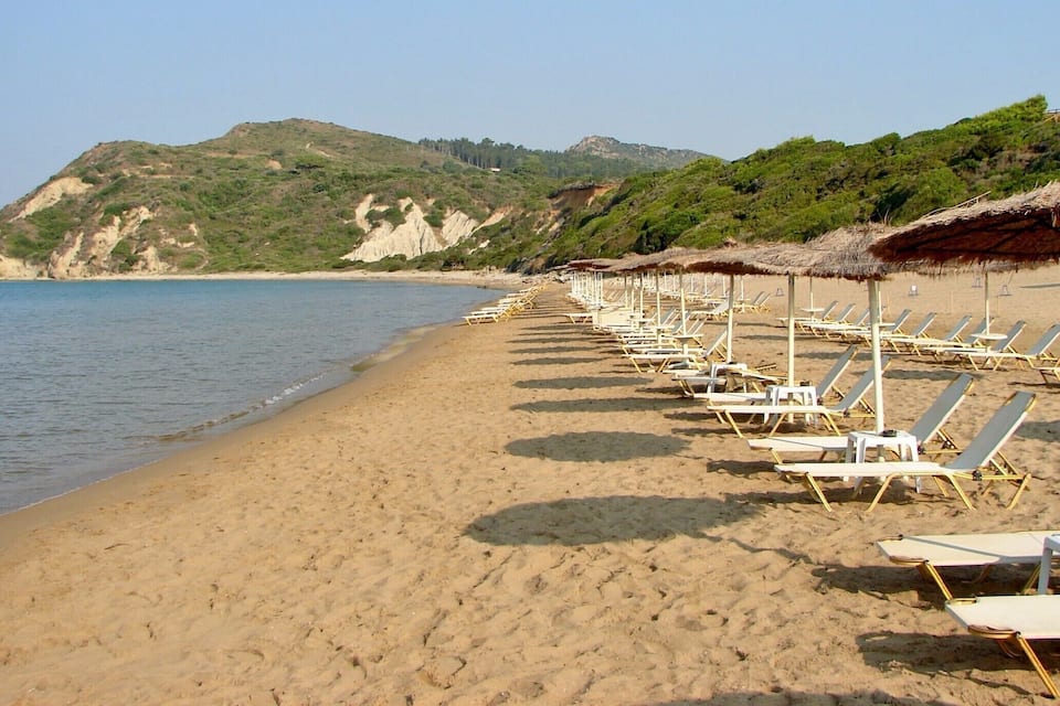 Uma praia com fileiras de espreguiçadeiras e guarda-sóis de palha, mar calmo e paisagem montanhosa ao fundo.
