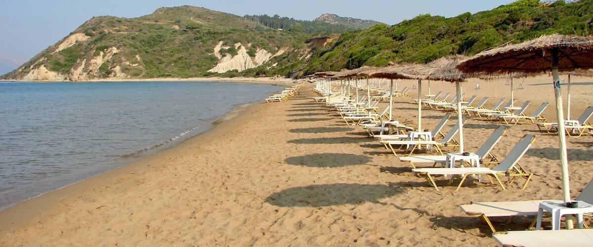 A beach with rows of sun loungers and thatched umbrellas, a calm sea, and a hilly landscape in the background.