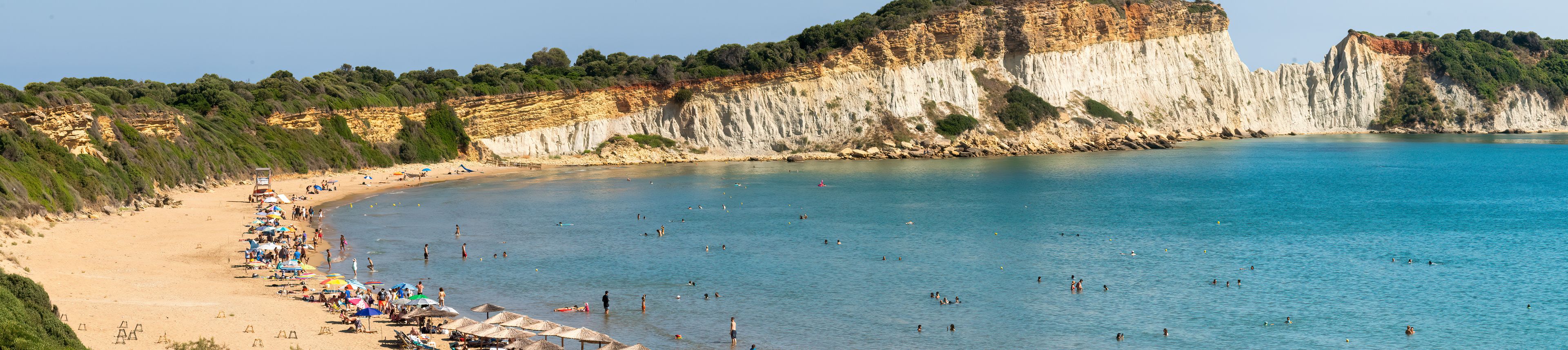 Une plage côtière aux eaux bleues et limpides, avec un rivage sablonneux et une falaise rocheuse couverte de végétation.