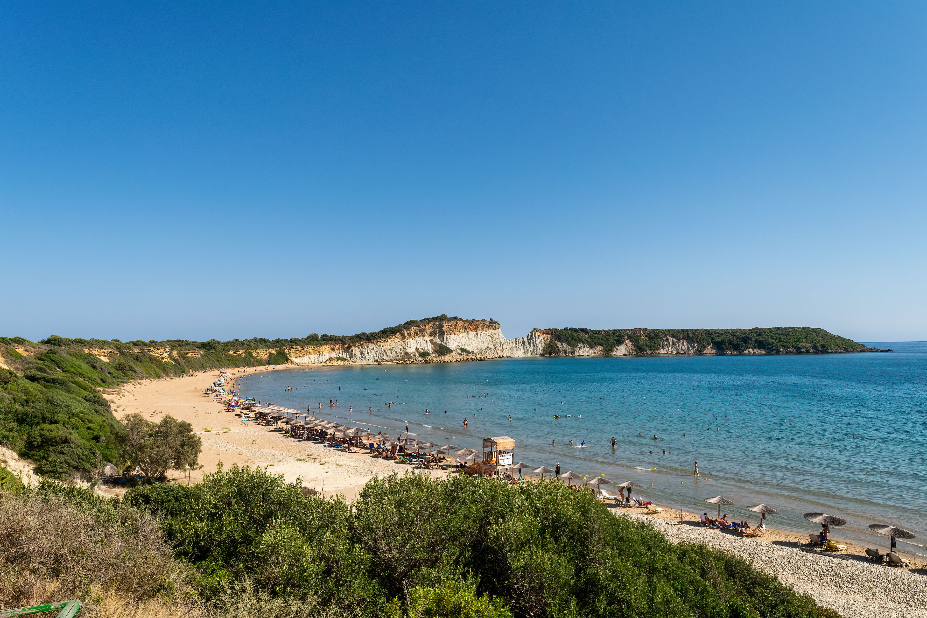 Une plage côtière aux eaux d’un bleu limpide, bordée de sable fin, avec une falaise imposante en arrière-plan.