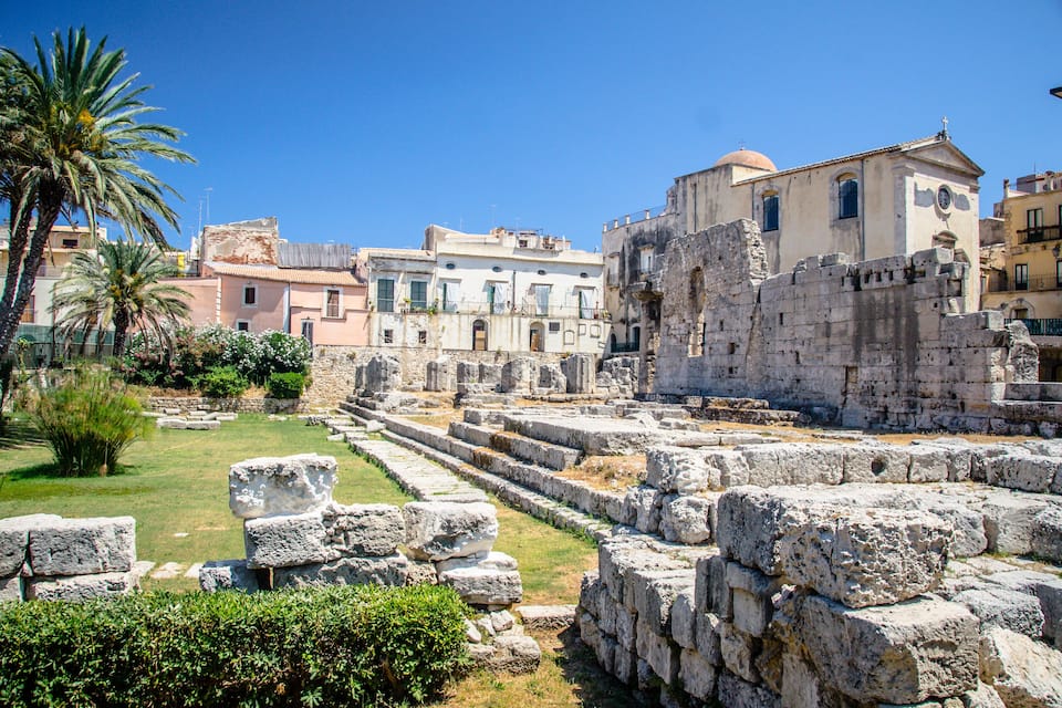 Ancient ruins with stone steps and walls, palm trees, and modern buildings in the background.
