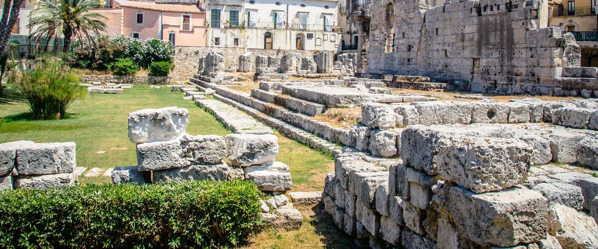 Ancient ruins with stone steps and walls, palm trees, and modern buildings in the background.
