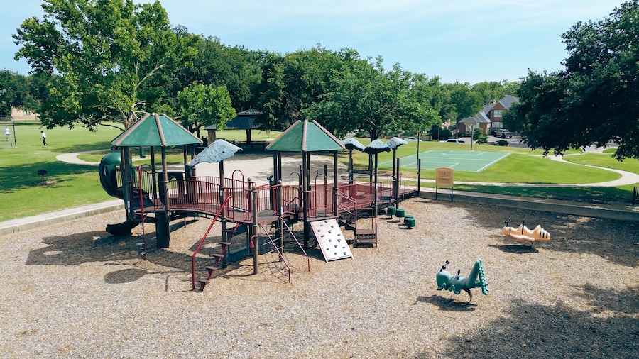 People playing baseball softball near backstop netting in grassy community park with modern playground equipment, hopper fun bouncer string rider, basketball court, upscale house, Plano, aerial