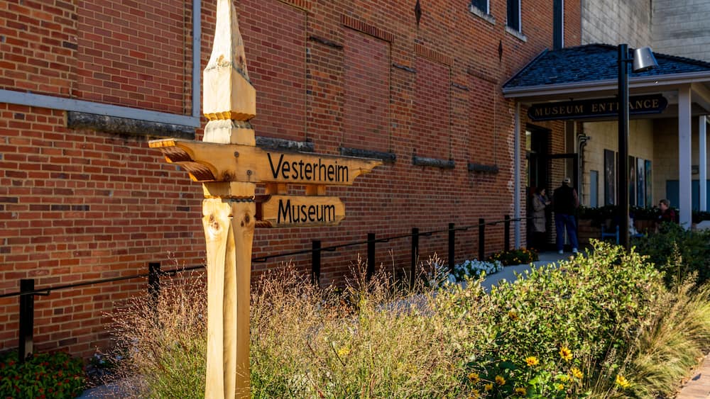 A wooden signpost with 'Vesterheim Museum' written on it, in front of a brick building with a sign reading 'Museum Entrance'.