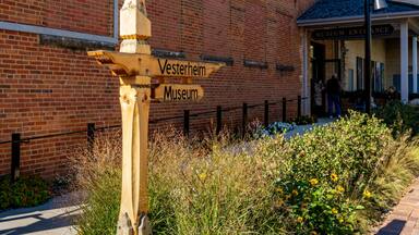 A wooden signpost with 'Vesterheim Museum' written on it, in front of a brick building with a sign reading 'Museum Entrance'.