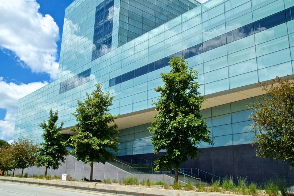 A modern glass building with a reflective facade, surrounded by trees and a clear blue sky.