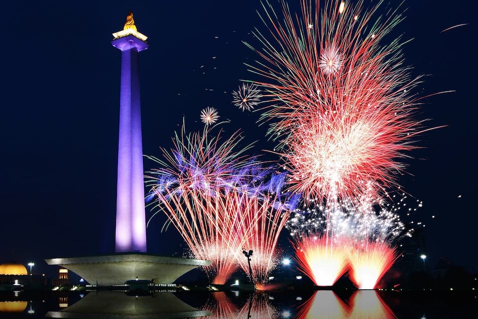 A night scene with a tall illuminated tower, fireworks, and a body of water reflecting the lights.