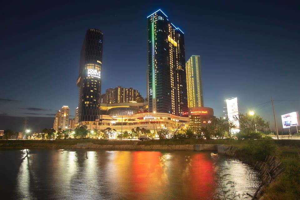 Un horizonte urbano nocturno con edificios iluminados, un río que refleja luces y un centro comercial con señalización visible.