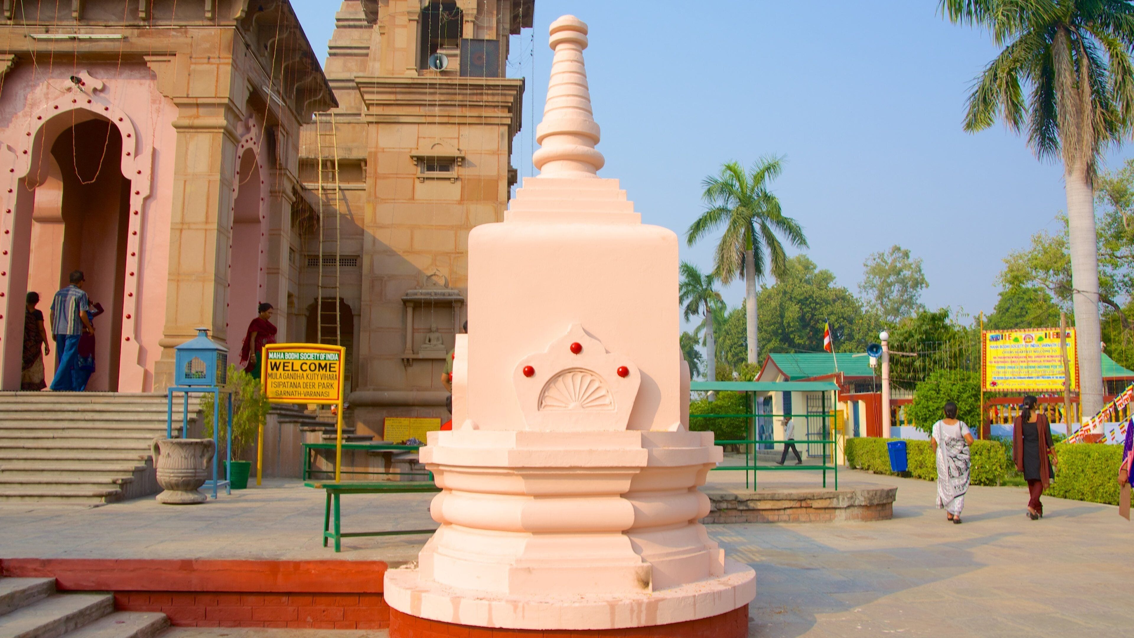 A large white stupa with a decorative top, surrounded by a red brick base. In the background, there is a pink temple with a signboard and a green building with a flag.