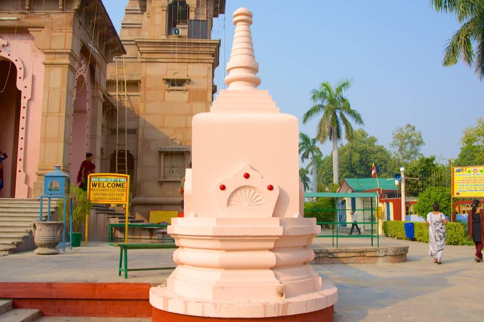 Una gran stupa blanca con un adorno en la parte superior, rodeada por una base de ladrillo rojo. En el fondo, se observa un templo rosa con un cartel y un edificio verde con una bandera.