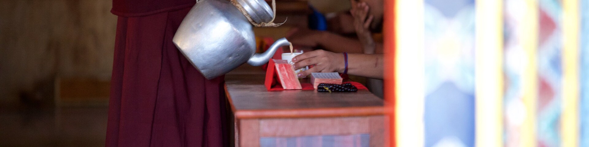 A person in a maroon robe pouring water from a metal container into a cup.