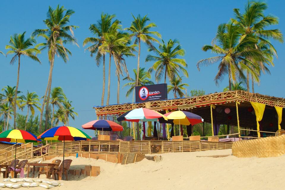 A beach with palm trees, a wooden structure with colorful umbrellas, and a sign for 'Garden Bar & Restaurant'.
