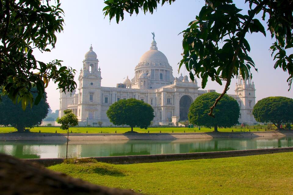 A grand building with domes and towers, surrounded by a calm water body and lush greenery.