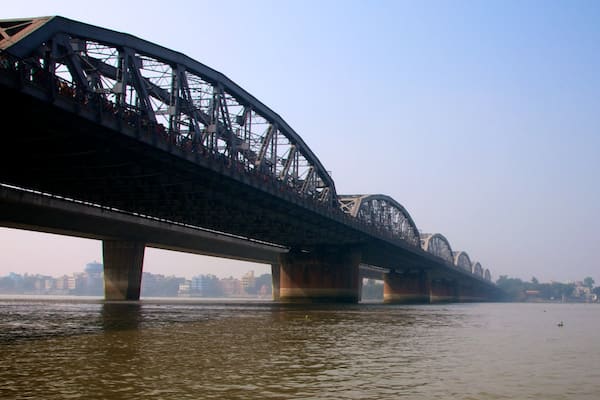 Eine lange Stahlbrücke überspannt einen Fluss, im Hintergrund eine Stadtlandschaft.