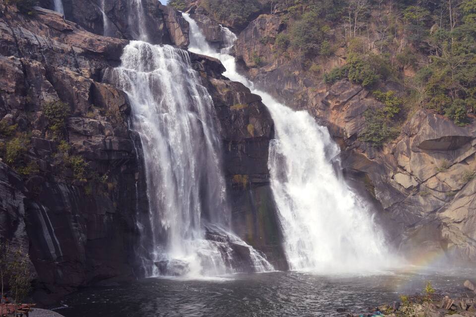 Une cascade à plusieurs niveaux dévalant des falaises rocheuses pour se jeter dans un bassin en contrebas, le tout entouré d’une forêt dense.