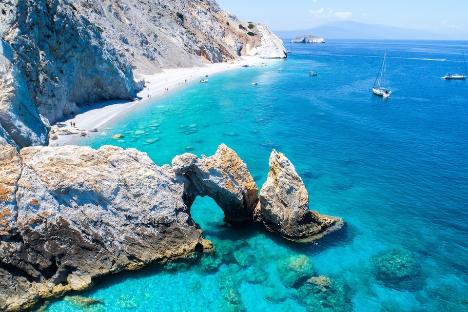 A coastal landscape with clear turquoise waters, rocky formations, and a sandy beach.