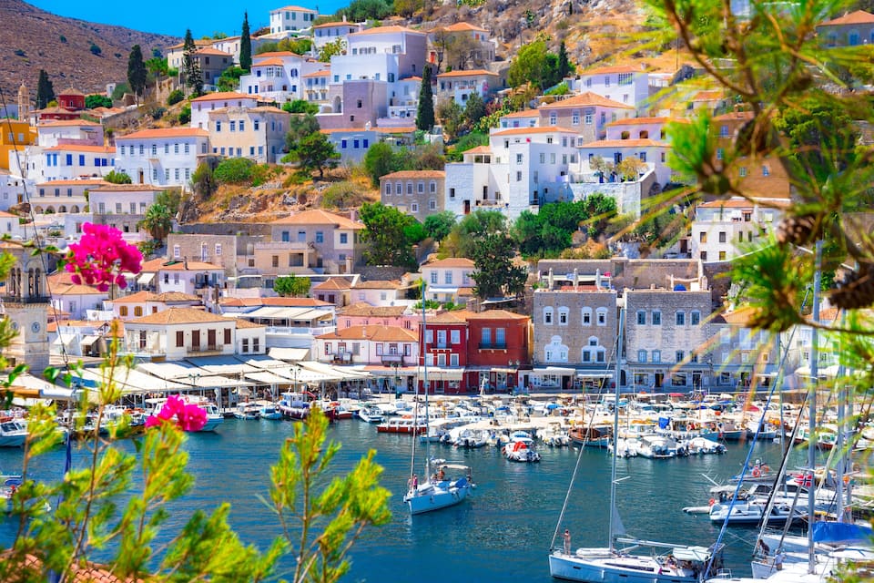 Un pueblo costero con edificios blancos, un puerto deportivo con barcos y un paisaje montañoso de fondo.
