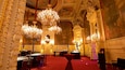 An opulent interior with ornate chandeliers, red carpet flooring, and classical wall paintings.