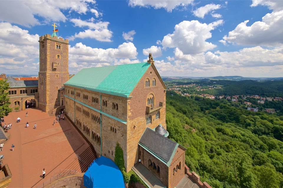 A historic church with a green roof and a tall tower, surrounded by lush greenery and a village in the background.