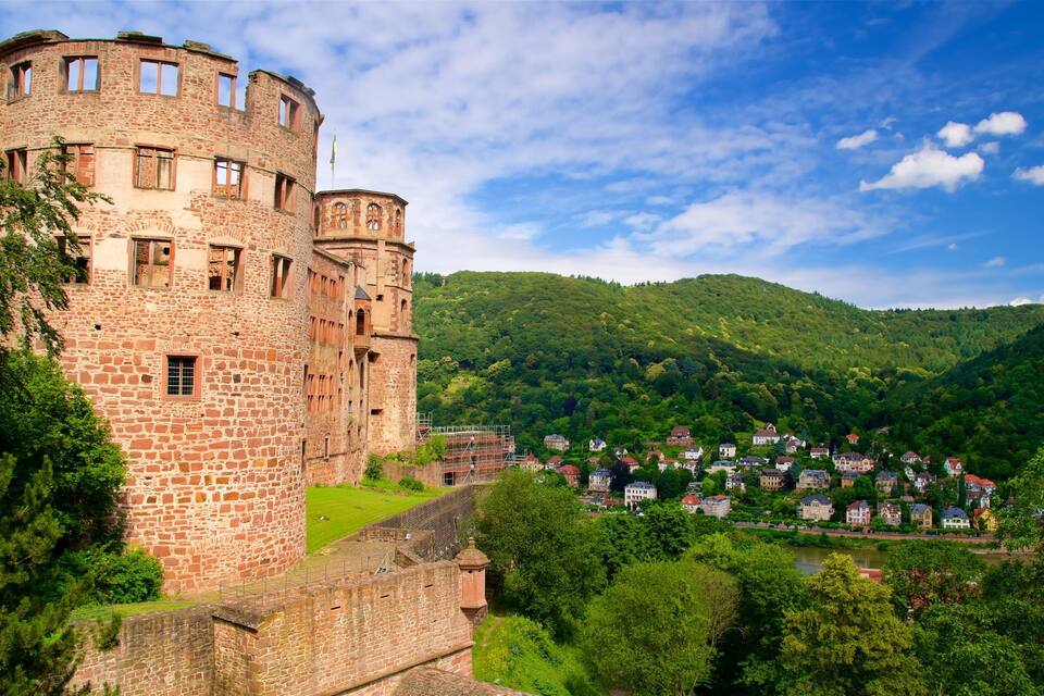 Um castelo histórico com uma torre redonda, rodeado por vegetação exuberante e uma aldeia no vale.