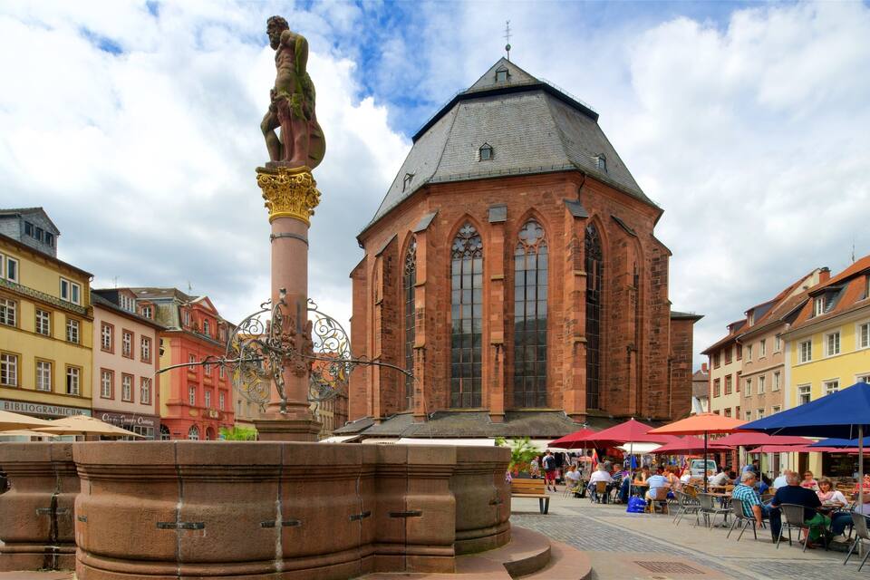 Ein historisches Gebäude mit spitzer Dachform und großen Fenstern, umgeben von farbenfrohen Gebäuden und einem öffentlichen Platz mit Menschen und Außenbestuhlung.