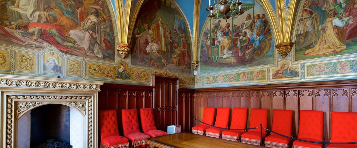 An ornate hall with red chairs, a long table, and elaborate frescoes on the ceiling.