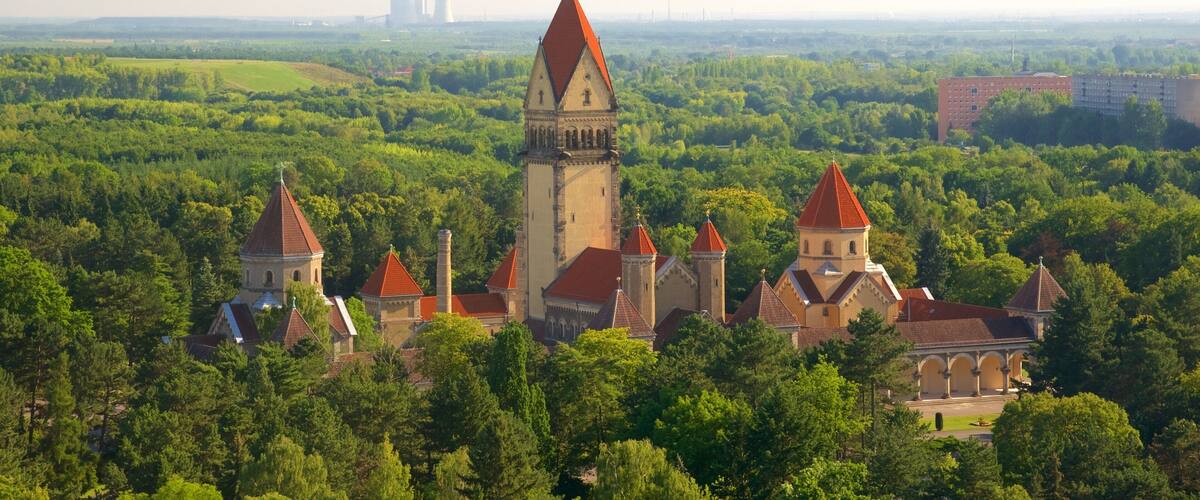 A castle complex with multiple towers and a church, surrounded by dense forest and a distant industrial facility.