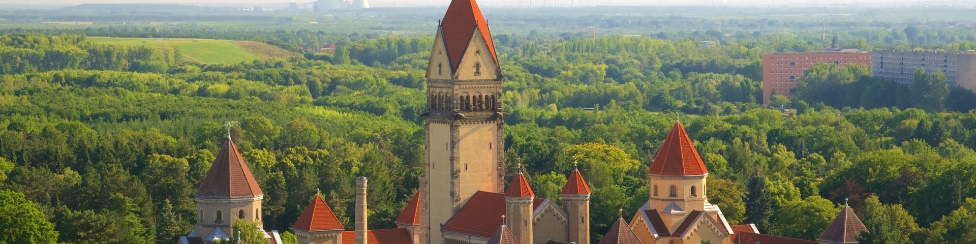 A castle complex with multiple towers and a church, surrounded by dense forest and a distant industrial facility.