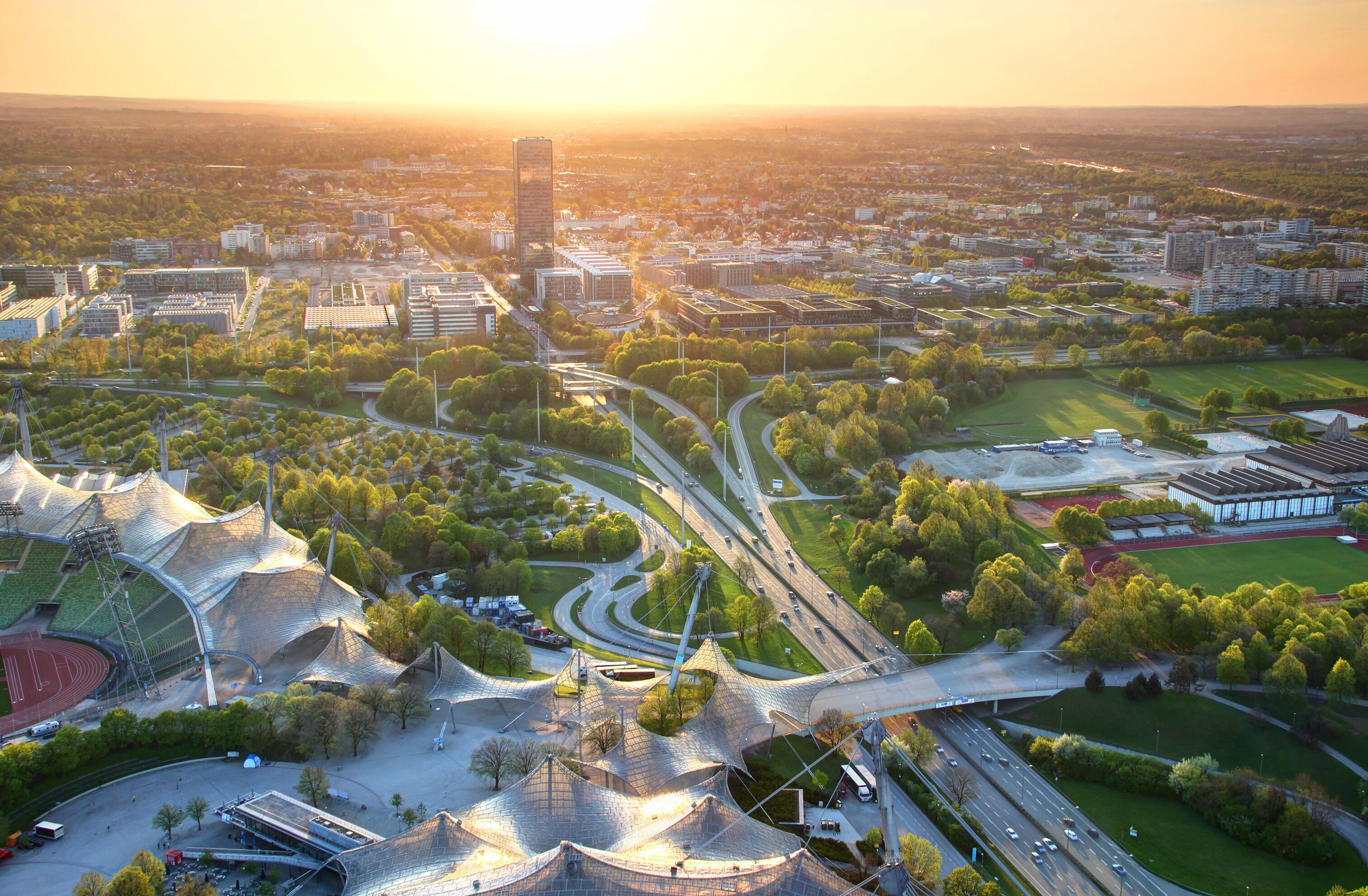 Modern European aerial cityscape with commercial and sports facilities, parks, multilane highway junction, tower blocks in outskirts illuminated by warm glowing sunlight at dusk Munchen Bayern Germany