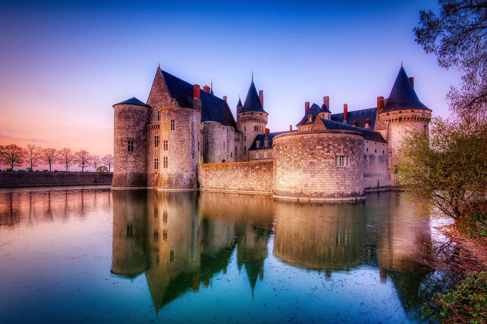 A historic castle with multiple towers and a moat, surrounded by trees and reflecting in calm water.