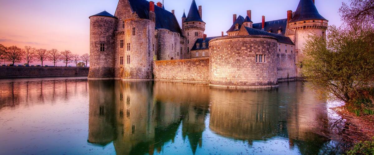 A historic castle with multiple towers and a moat, surrounded by trees and reflecting in calm water.